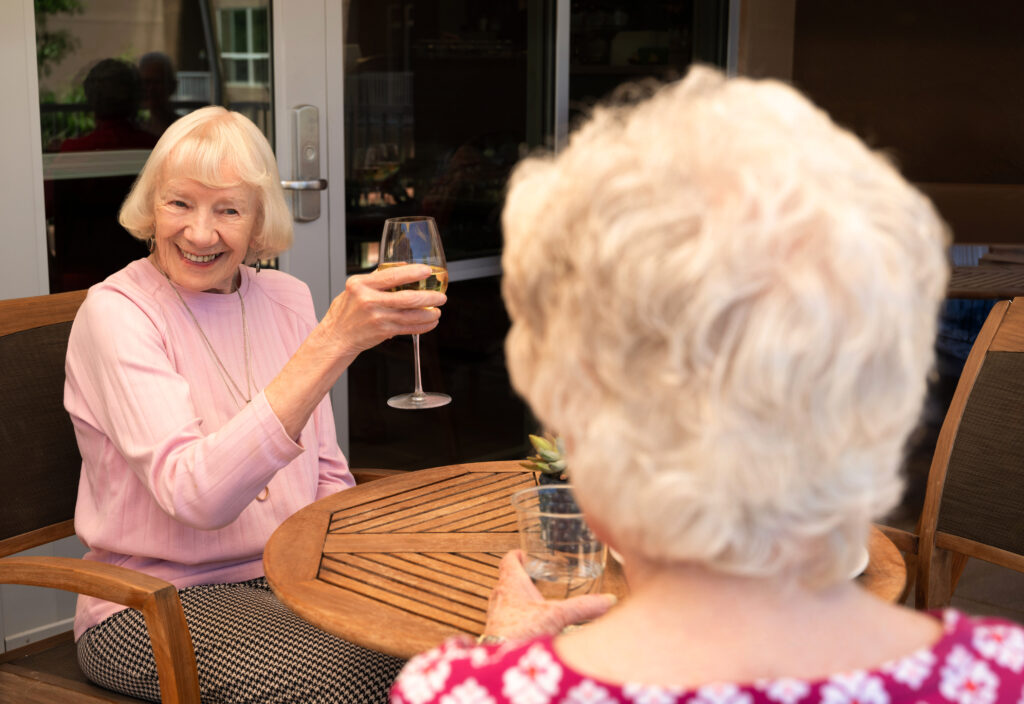 Two older women seated at a round wooden patio table outdoors raising glasses of white wine in a toast, one facing the camera.